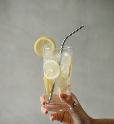 ruka drží sklenený pohár so slamkou, citrónovou vodou a neošúpanými plátkami citrónu
